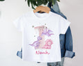 White toddler t-shirt with a pink number 1 and dinosaur illustrations. The design features three cartoon dinosaurs in purple and pink, with personalised name in pink script underneath.