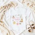 Personalised White toddler t-shirt featuring an Easter design with a bunny, chick, teddy bears, and the text "MY FIRST EASTER 2026". Cream-coloured shoes are placed beside the shirt. The design is in pastel colours.