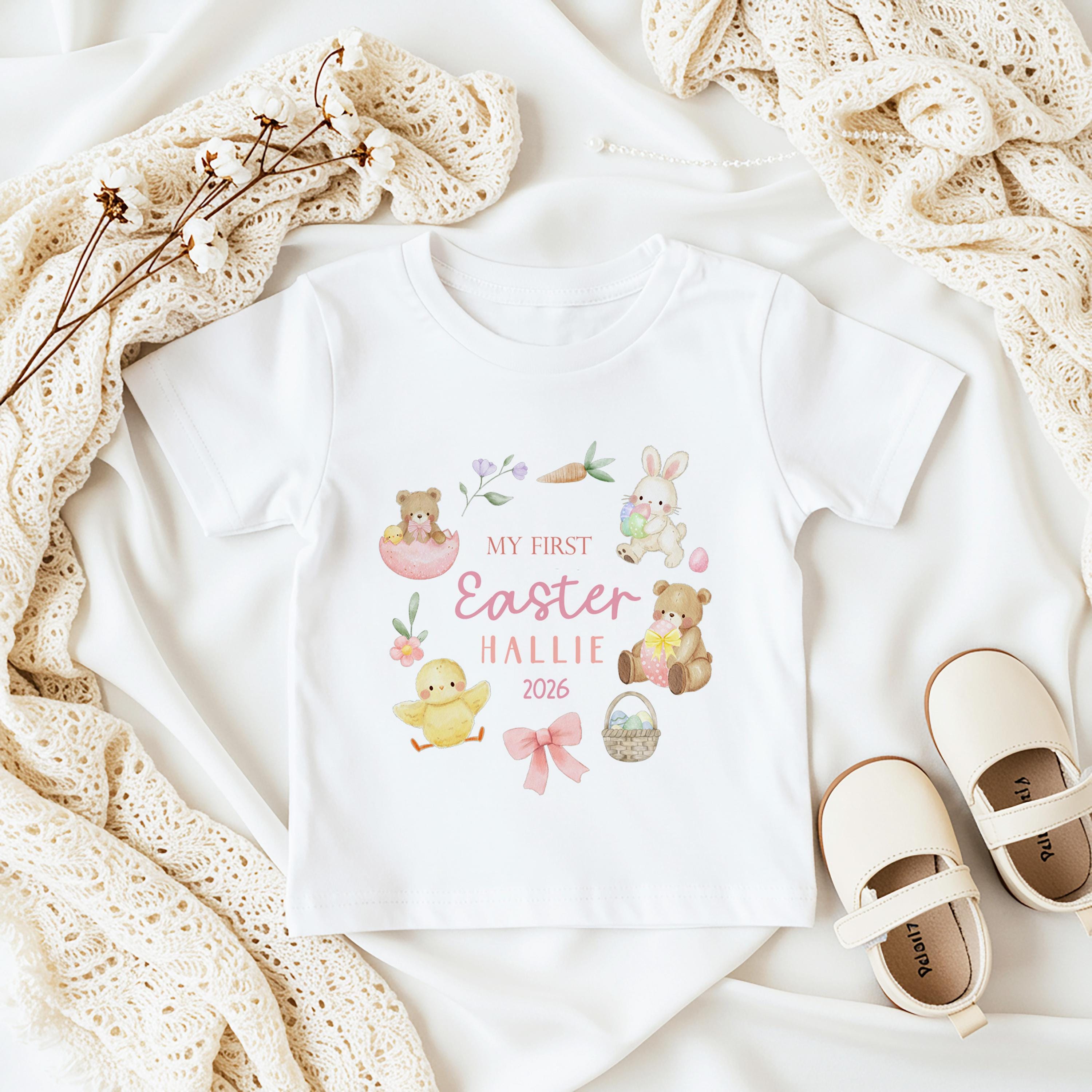 Personalised White toddler t-shirt featuring an Easter design with a bunny, chick, teddy bears, and the text "MY FIRST EASTER 2026". Cream-coloured shoes are placed beside the shirt. The design is in pastel colours.